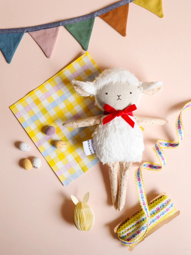 Handmade lamb doll with a fluffy white body, stitched face, beige floral fabric arms and legs, and a small red velvet bow tied around its neck.