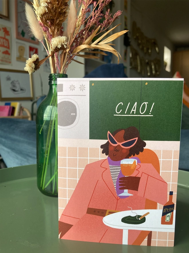 Greetings card illustrated with someone in their kitchen and an Aperol Spritz. The text says 'Ciao'. Placed on a table with flowers in the background.
