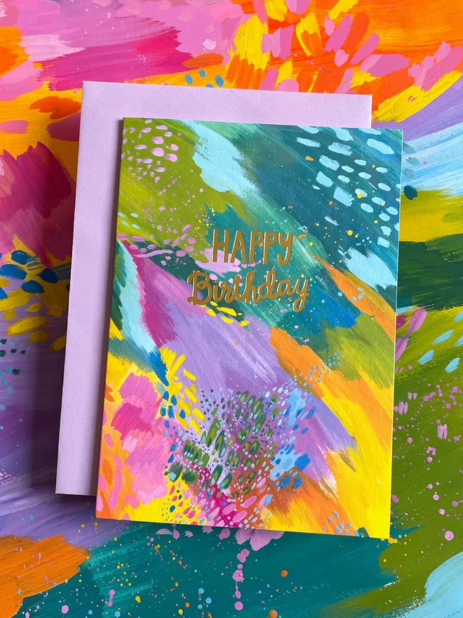 The colourful Raspberry Blossom birthday card sits on top of a lilac envelope and the painting used to create its vibrant design