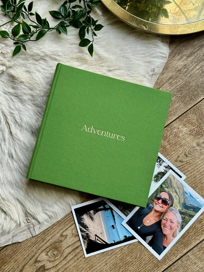 A hand-bound green photo album printed with the words 'Adventures' in gold lettering rests on a bookshelf next to rows of red books.