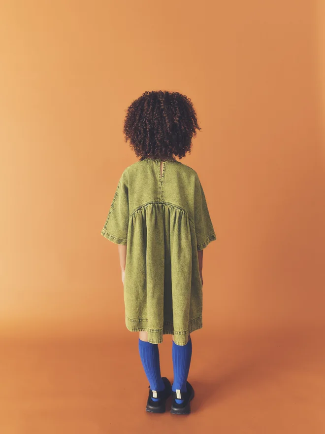 Back view of a child against a burnt orange backdrop, showing the gathered back and keyhole closure of the Studio Koter Rumi dress in green acid-wash denim.