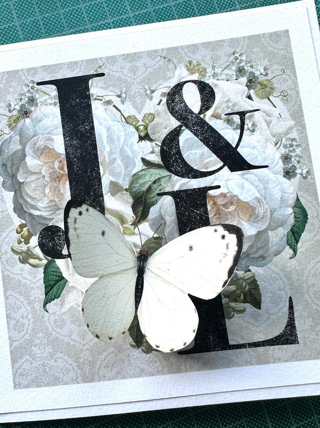 A handmade Diamond Wedding anniversary card featuring two initials of a couples names, placed inside a heart design with vintage white roses and on a taupe background. The recipients initials are hand written on the front of card and hand signed, A white 3D papercut butterfly sits on the heart and wings can be folded out for display.