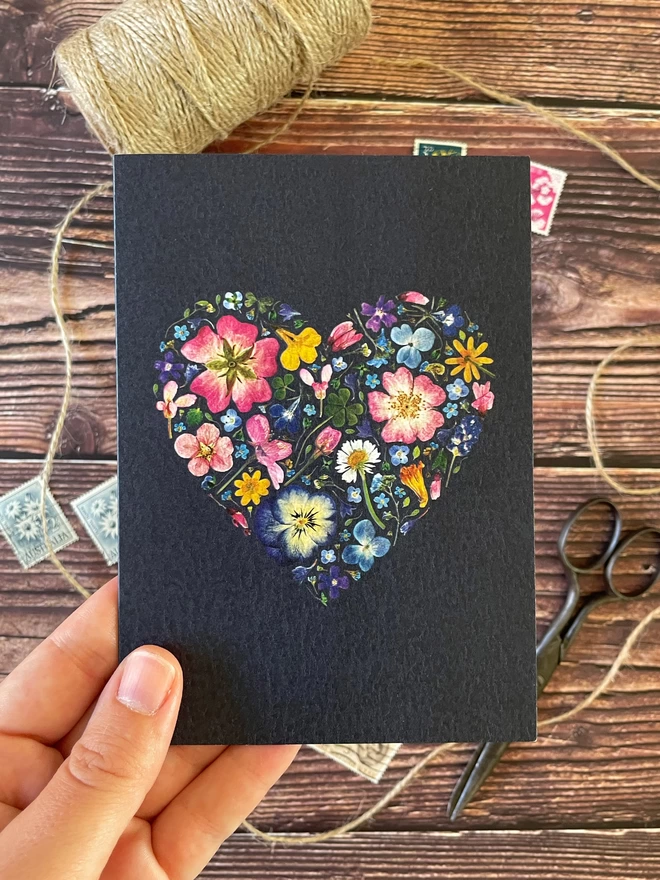 Hand Holding Greetings Card with Pressed Flower Heart Design on Dark Blue-Black Background - Wooden Desktop with Brown String, Vintage Scissors, Floral Postage Stamps