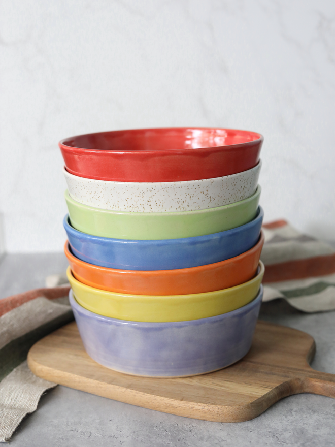 A neatly stacked set of seven glazed ceramic pasta bowls in rainbow colours — red, speckled white, green, blue, orange, yellow, and lavender — on a wooden chopping board, surrounded by linen napkins, against a pale grey background.