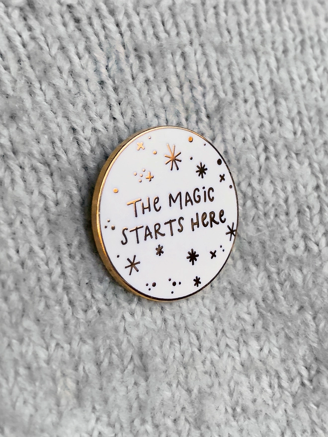 A white and gold enamel pin badge with a starry design and the words "The magic starts here" is pinned to a grey jumper.