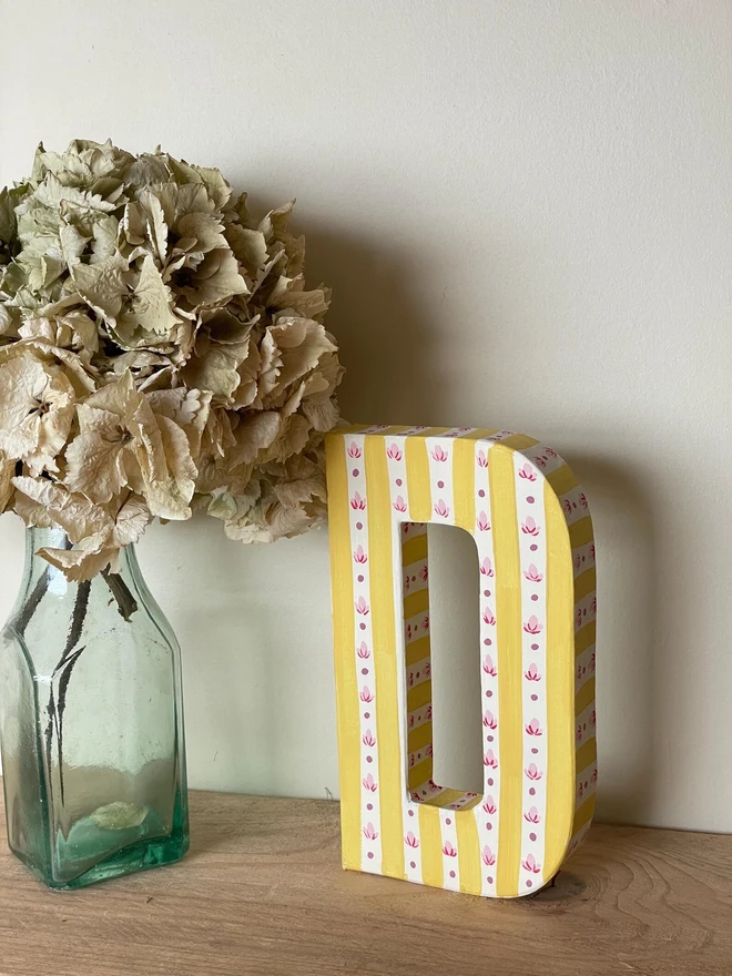 A hand-painted decorative initial letter  with stripes on a wooden table against a light wall. 