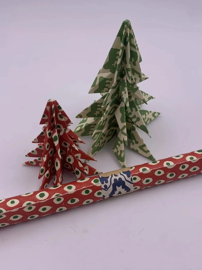 Christmas Tree Kits. Two origami Christmas trees folded from block-printed paper, one in red with green and cream spots and a taller one in green and cream leaf print. The layered paper trees make a handmade festive table or mantelpiece decoration.
