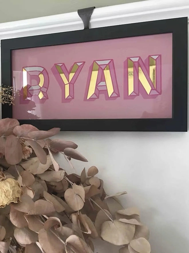 Personalised Name Wall Plaques, a pink personalised gold leaf sign sitting in a black frame hanging in front of a white wall. 