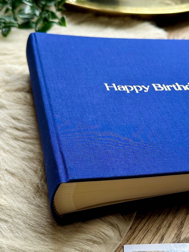 a close up image of a photo album with the words Happy Birthday on the front. The photo album is blue and made from linen, the printing on the front is gold. It has been placed on a sheepskin rug.
