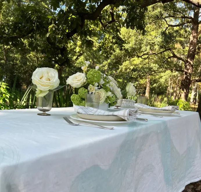 Imola Tablecloth. Light blue and white tablecloth with subtle abstract patterns, styled on an outdoor dining table with plates, glassware, and white floral arrangements.