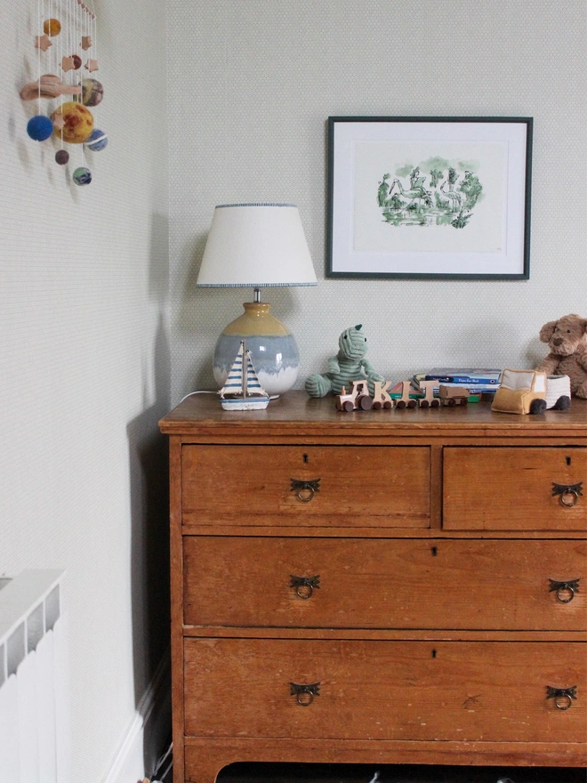 Cream empire lampshade with blue striped trim on a ceramic table lamp, styled on an antique wooden chest of drawers in a children’s bedroom with wooden toys, soft toys and framed artwork.