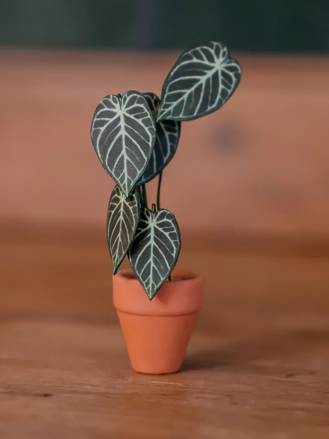 A miniature Anthurium Crystal Hope paper plant ornament sat on a grey shelf amongst some real plants