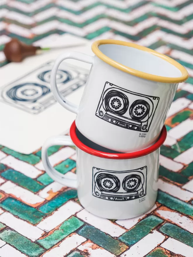 Picture of 2 White Enamel Mugs with a cassette tape design etched onto it, taken from an original Lino Print. 1 has a yellow rim, and the other has a red rim.