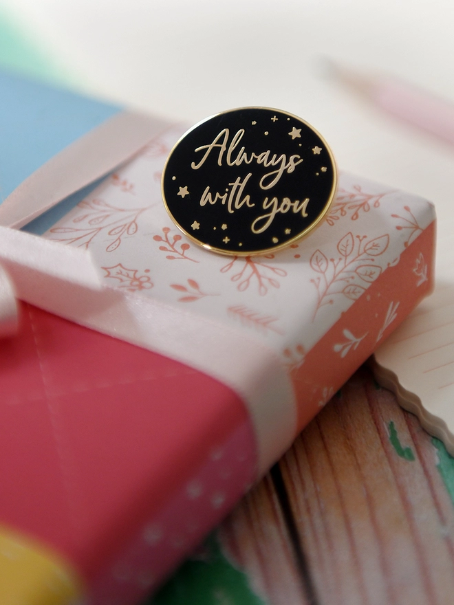 A black and gold enamel pin badge rests on a wrapped gift. It has a subtle star design and the words "Always with you".