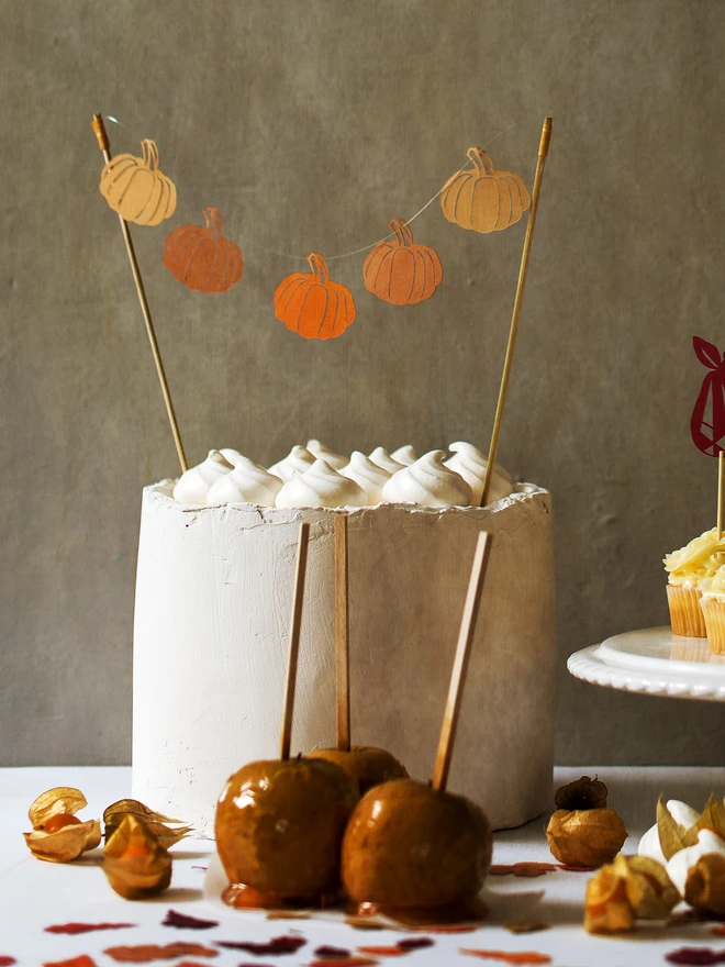 Cake Topper with 5 paper cutout pumpkins displayed on a plain white cake. Cake is placed on a table with toffee apples, cupcakes and paper leaf confetti