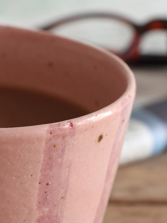 Close-up shot of a Handmade Ceramic Stoneware Mug in Pink Stripe. A newspaper and a pair of glasses can be seen in the background. 