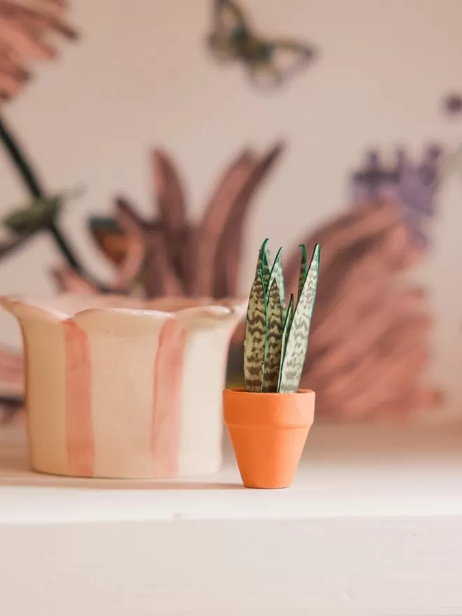 A miniature replica Sanseveria Snake Plant paper plant ornament in a terracotta pot sat on a wooden log slice with two other paper plants to the left of it (an Anthurium Regale and an Anthurium Clarinervium)