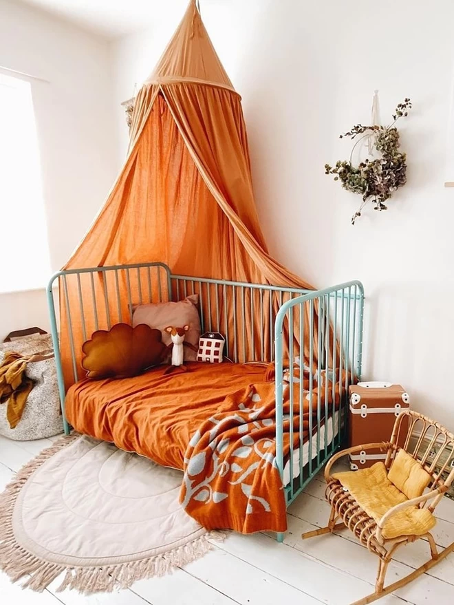 A warm and inviting children's bedroom showing a turquoise iron cot with the side dropped down. A rust orange canopy hangs above the cot and the bed is made with a rust orange sheet and the briar rose junior blanket draped at the end. A small wicked rocking chair sits at the end of the bed and a plant hangs on the wall.