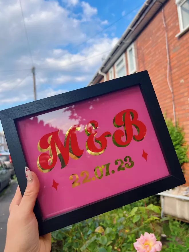 Black framed 'M & B' wedding sign with a curly script style on pink background, with red lettering and gold leaf shade and date.