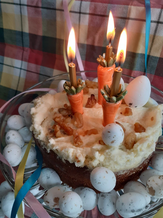 Carrot cake decorated with handmade ceramic carrot candleholders and lit birthday candles, surrounded by speckled Easter eggs on a glass cake stand.