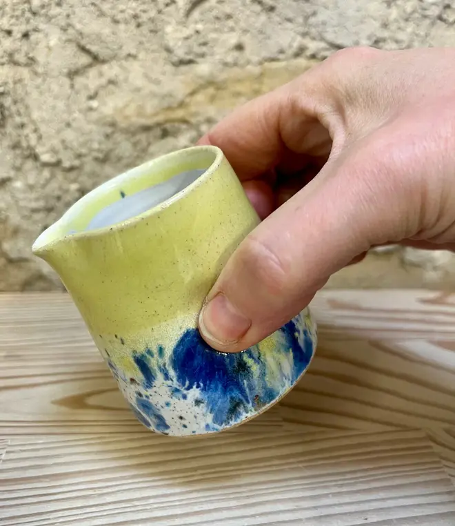 Shoreline Tiny Jug, a colourful tiny jug placed on a wooden surface. It sits in front of a brick wall backdrop. In the image its being poured. 