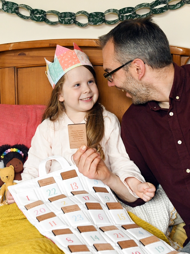 A young girl sits on a bed beside her father as they read a wooden story card, and a fabric advent calendar rests on their laps.