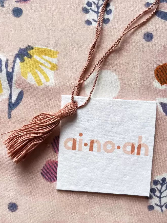 detail of recycled white tag with ainhoa logo and dusty pink tassel hanging loop over a printed pattern