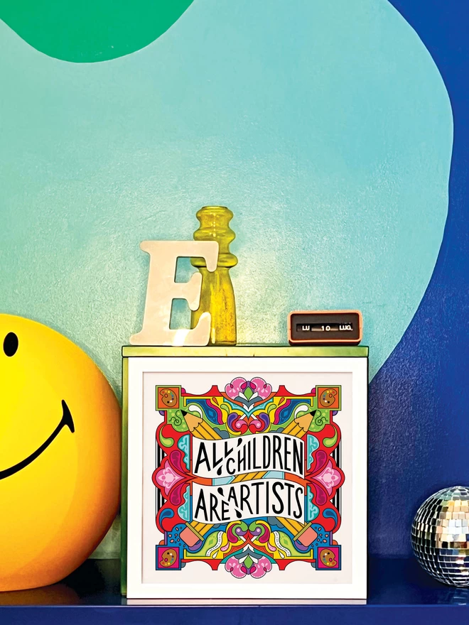 A white frame surrounds the print, which has the words All Children are Artist at the centre of this multi-coloured vibrant illustration which includes a cross of pencils, and artists palettes the corners, which is propped against a turquoise and dark blue wall. Next to the frame is a disco ball, a letter ‘E’ ornament, a yellow glass vase, an orange Italian plastic calendar showing the date as ‘LU 10 LUG’ and a large light up yellow Smiley lamp.