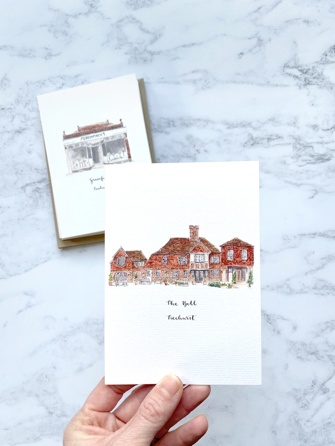 Greetings card showing The Bell, in Ticehurst, a beautiful brick building with black and white framed window and door which is open welcoming you into the shop. The card is held by a white womans hand between finger and thumb. The watercolour style is painted with a black pen outline and organic loose style with small details. The card sits on the top of a pack of cards with a white paper band around.