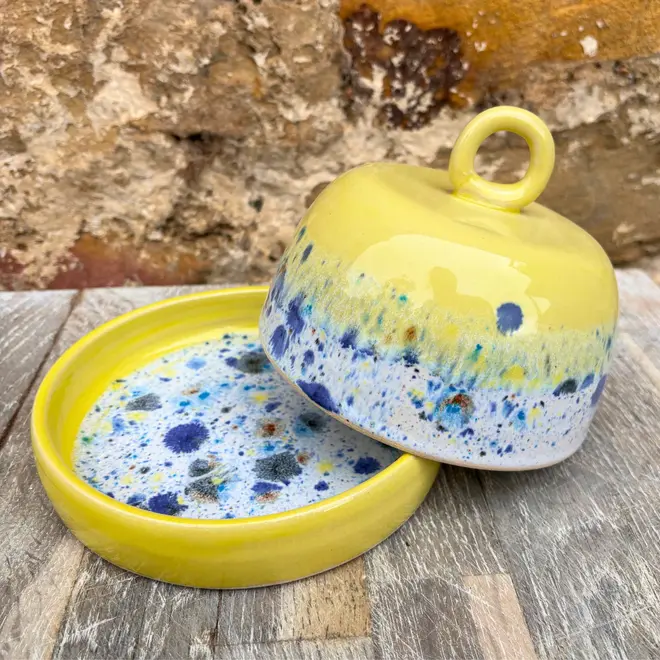 Shoreline Butter Dish, a colourful butter dish placed on a wooden surface against a brick wall. The lid is coming off in the image. 