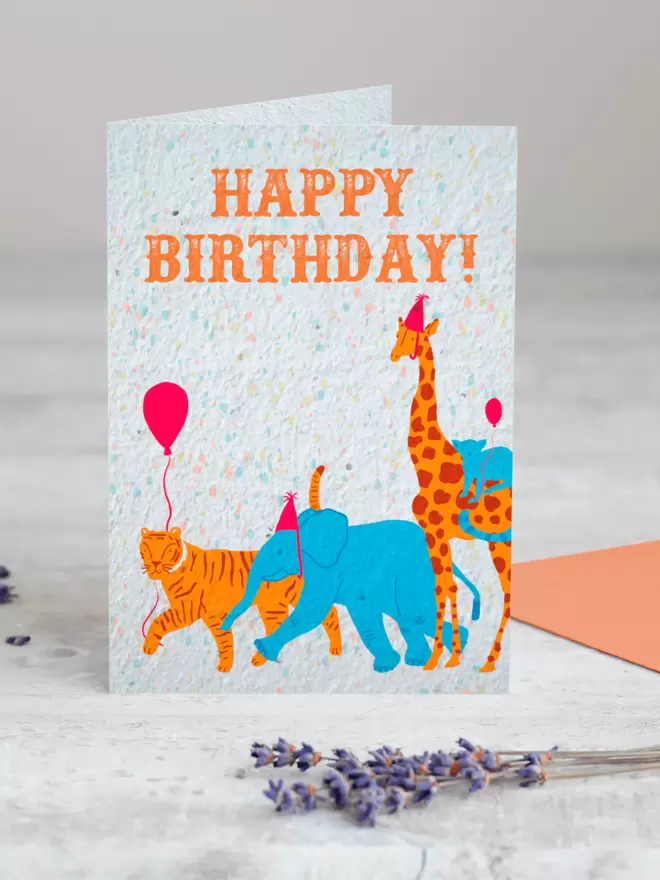 Plantable Card with Happy birthday and an illustration of an elephant and giraffe wearing party hats and a tiger and monkey each carrying a balloon with a sprig Lavender in the foreground