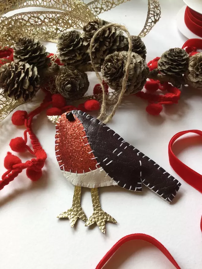 A leatherette robin Christmas tree decoration, hand stitched, with red glittery breast, sits amongst pine cones, red ribbon, and pom pom trim,