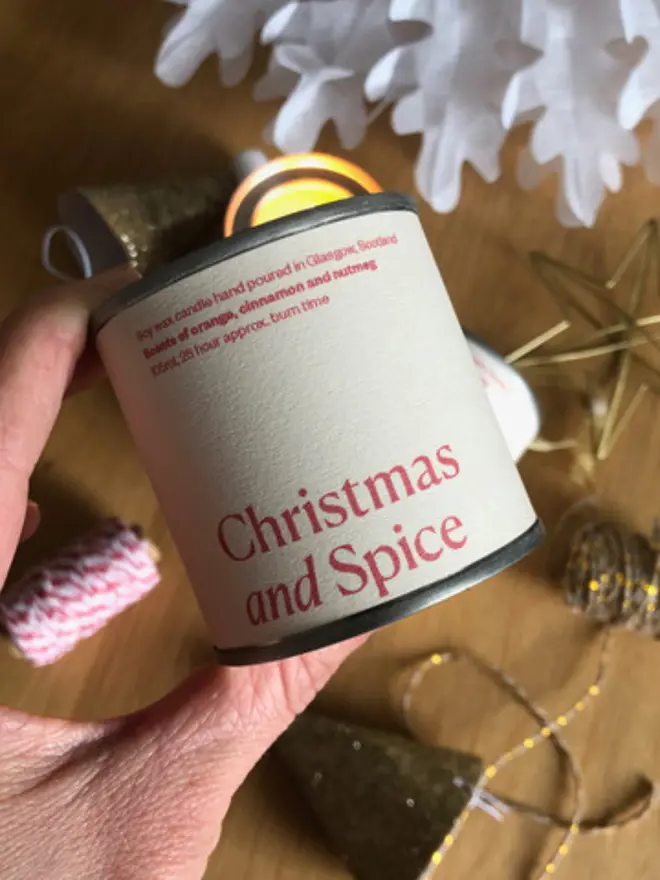 Christmas and Spice Wax Candle - a pale pink and red tin in a small and big size held up against some christmas items 