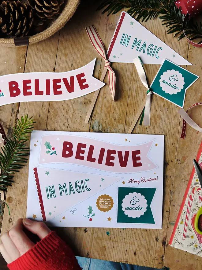 A Christmas card featuring three Christmas flags that read "Believe in magic & wonder" lays on a wooden table with various Christmas greenery, books & decorations around it.