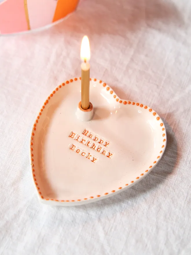Personalised ceramic heart birthday dish with a candle holder with an orange dotted rim design, placed on a plain white tablecloth 