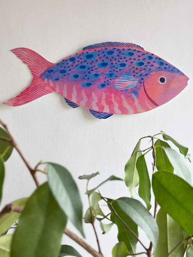 large wooden fish wall decoration. wooden fish decoration hung on white wall above houseplant. 