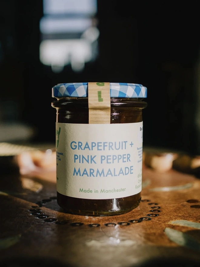 Jar Of Grapefruit & Pink Peppercorn Marmalade, jar of marmalade with a blue lid on a decorated plate on a table. 