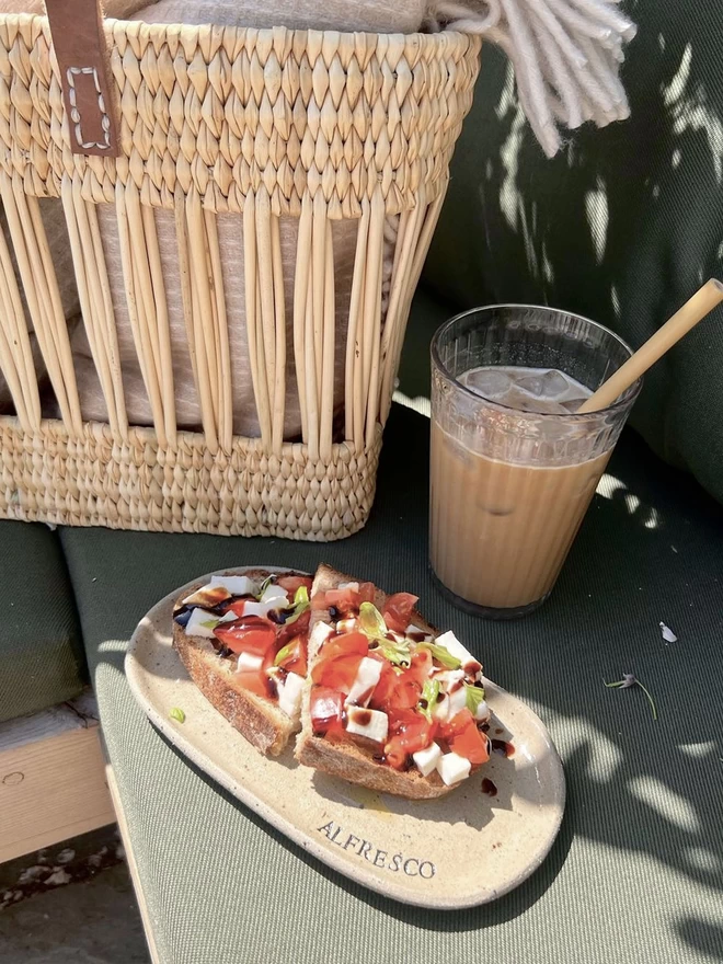beige, oval plate holding bruschetta beside a glass of iced coffee and a woven basket