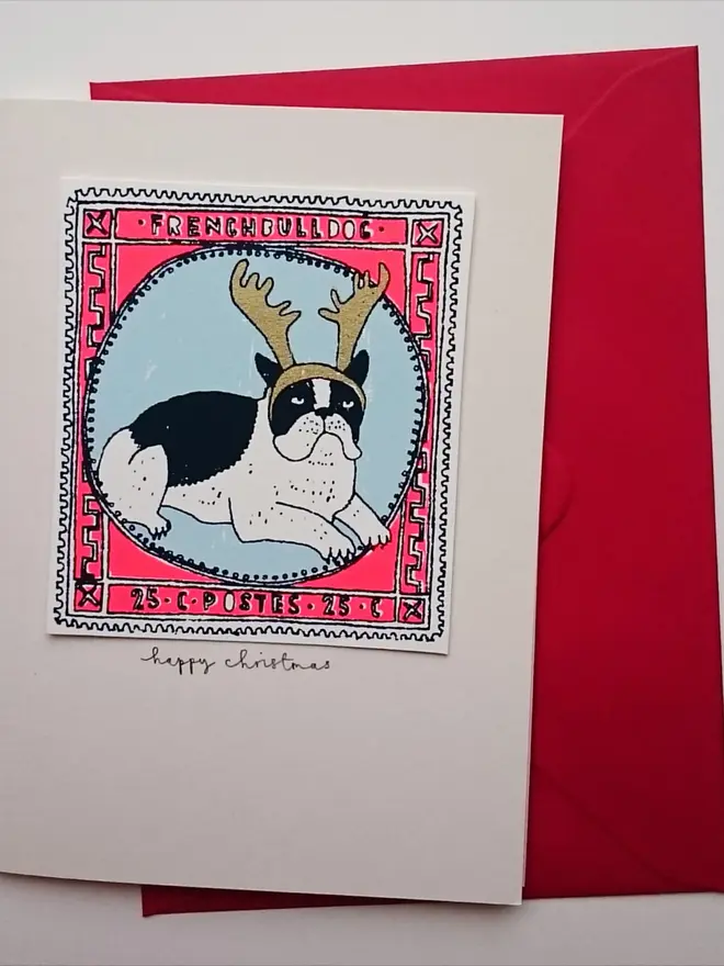 Happy Christmas French Bulldog Card, a white card with a colourful dog design in the centre on a white surface. This is placed above a red envelope. 