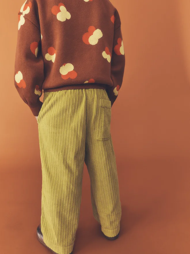 Close crop of a child with eyes closed against a burnt orange backdrop, wearing a Studio Koter chestnut jacquard jumper with chartreuse corduroy trousers.