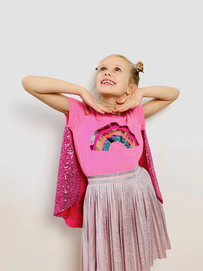 The front of a young girl wearing the big sister cape. The front of the cape hangs openly and is made from pink sequins.