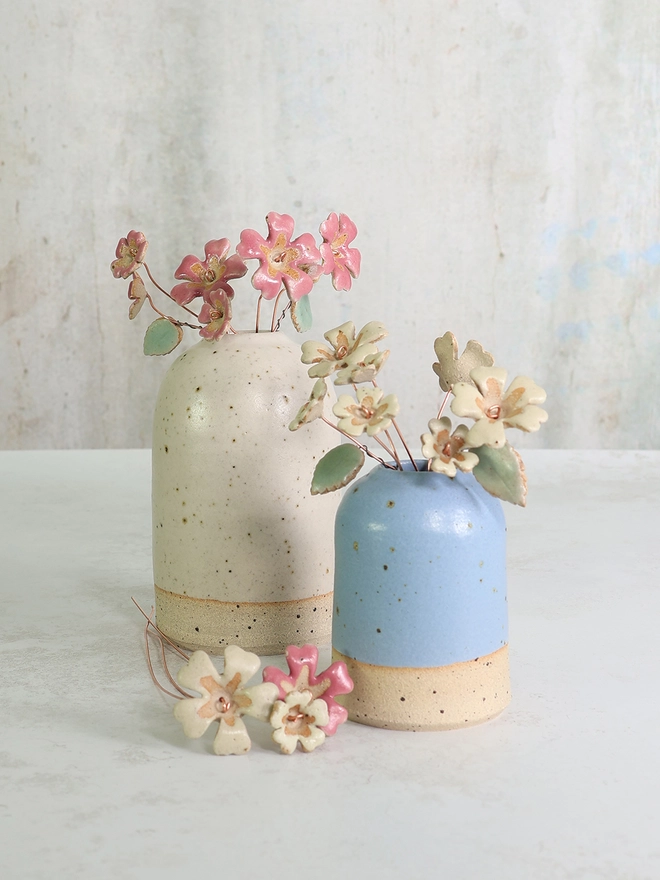 two handmade ceramic vases sit on a marble table top the smaller blue vase is in front and holds a posy of ceramic primroses the large white vase has a display of pink ceramic flowers in 