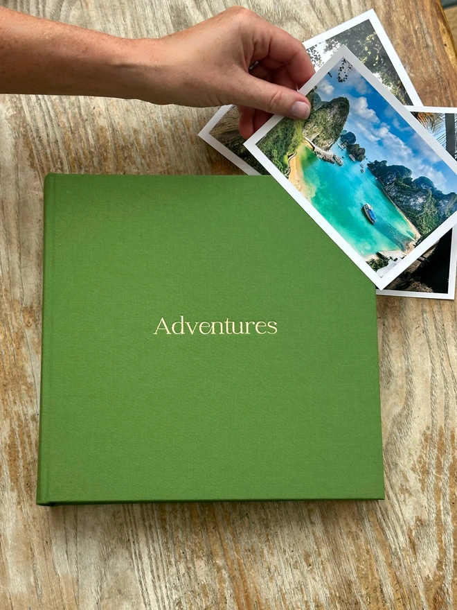 a green photo album lies on a wooden table. A person's arm is leaning over holding a photo of a sea and sand landscape.