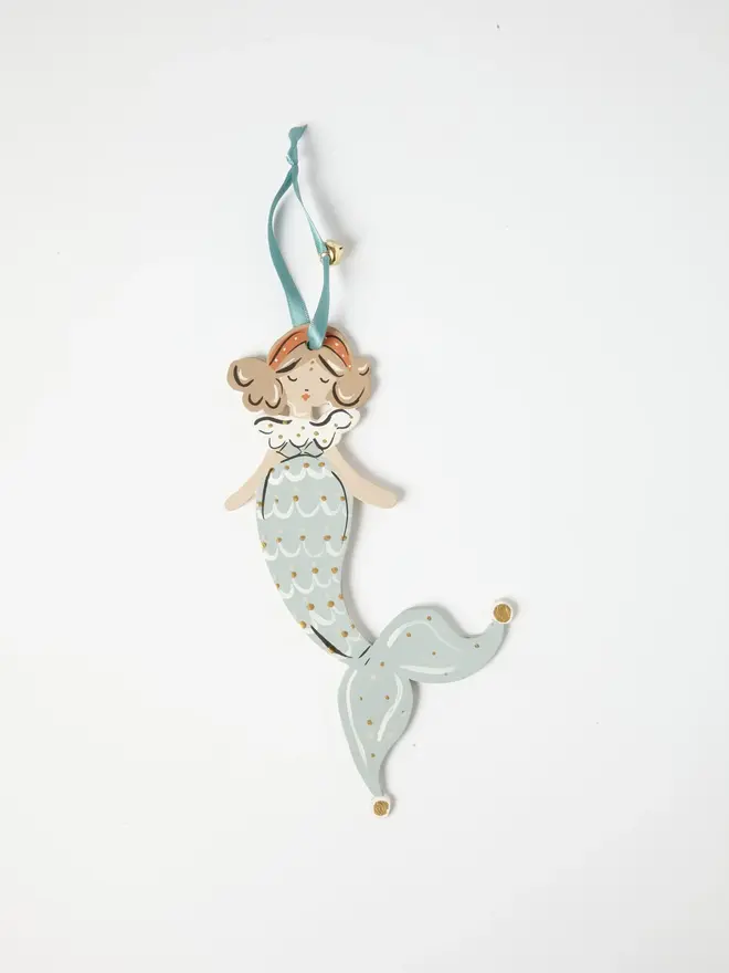 Mermaid Tree Dancer. a flat lay product shot of the mermaid tree dancer hanging ornament on a plain white background, showing a mermaid figure with curled hair and an orange headband, dressed in a pale blue scalloped scale-patterned tail with gold dot detailing. the tail curls into a jester-style pointed tip with gold-tipped ends, and the figure hangs from a teal ribbon with a small gold bell attached. hand-painted detailing gives the ornament a folk-art, storybook charm.