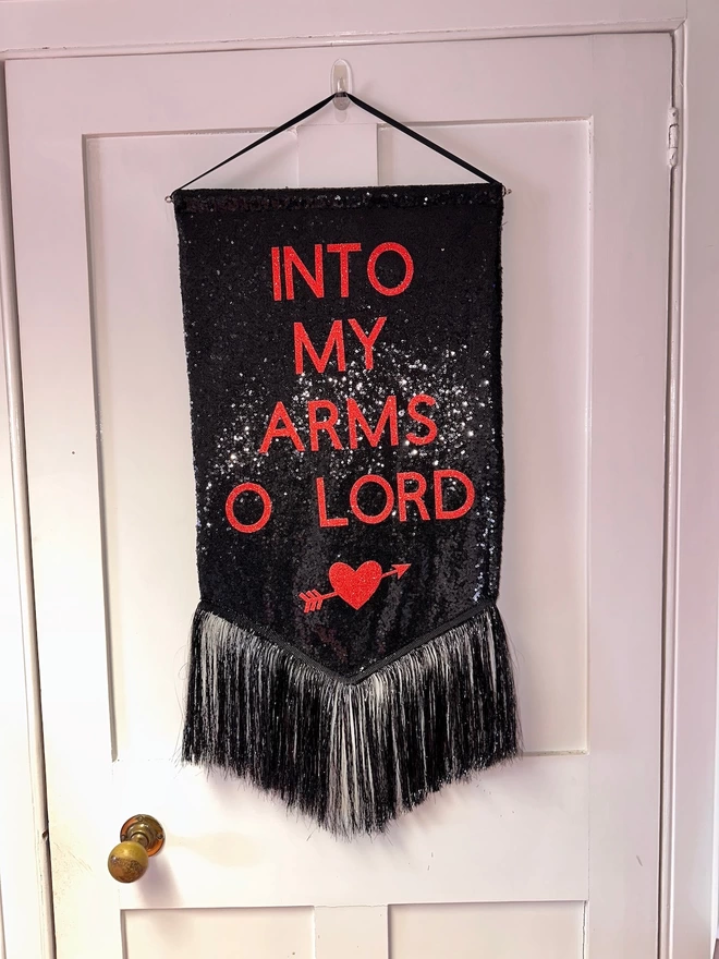 A black sequin banner with 'INTO MY ARS O LORD' and a heart features in red. It hangs on a black ribbon and has a black tinsel trim along the bottom.
