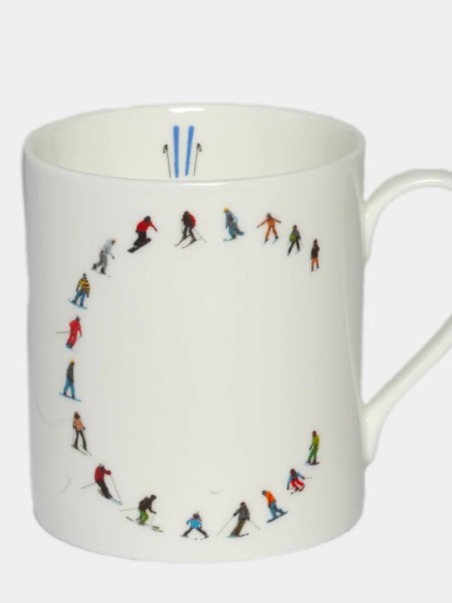 A simple white mug illustrated with a ‘C’ made of small, multicoloured skiers. The mug is set against a plain, white background.