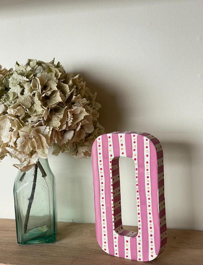A hand-painted decorative initial letter  with stripes on a wooden table against a light wall. 
