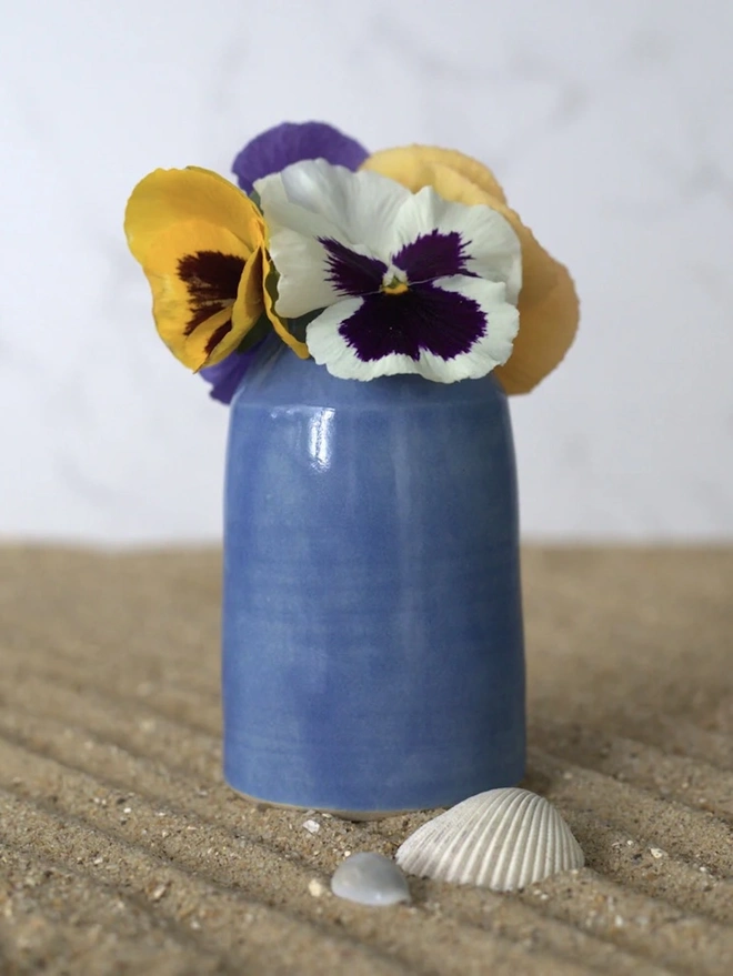 Classic Bud Vase, a colourful ceramic vase sitting on a plain surface against a grey backdrop. It is accompanied by some flowers and shells. 