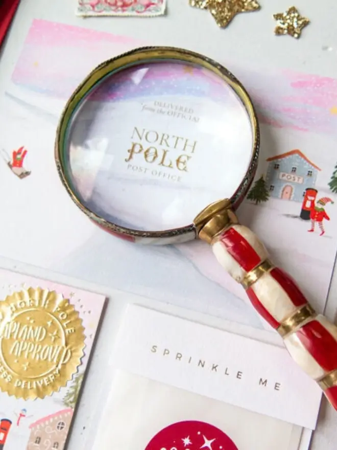 A candy-striped magnifying glass over a pink North Pole Post Office card with gold foil lettering.
