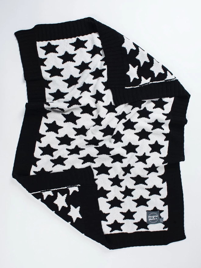 A flatlay of a knitted baby blanket with black and white star pattern and balck ribbed trim. The corners are slightly ruffled and turned back to reveal the reverse colourway.